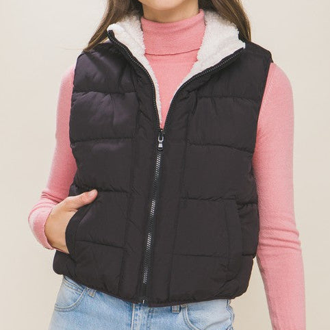 Woman wearing a black puffer vest over a pink sweater with a beige background